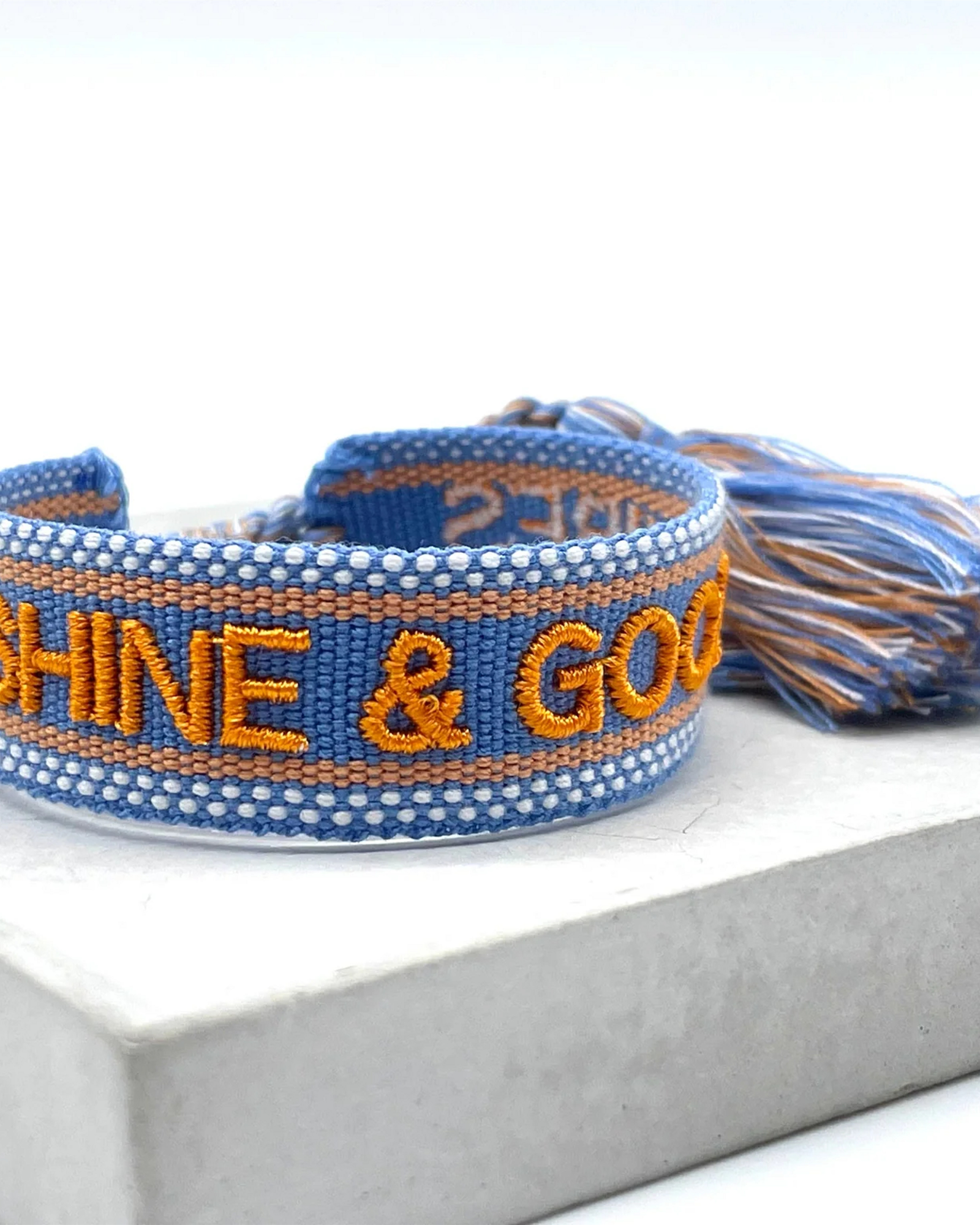 Close-up of adjustable blue and orange woven friendship bracelet featuring embroidered "SUNSHINE & GOOD VIBES" lettering and sliding knot tassel cords.