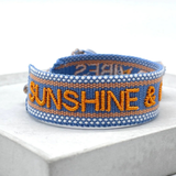 Close-up of adjustable blue and orange woven friendship bracelet featuring embroidered "SUNSHINE & GOOD VIBES" lettering and sliding knot tassel cords.
