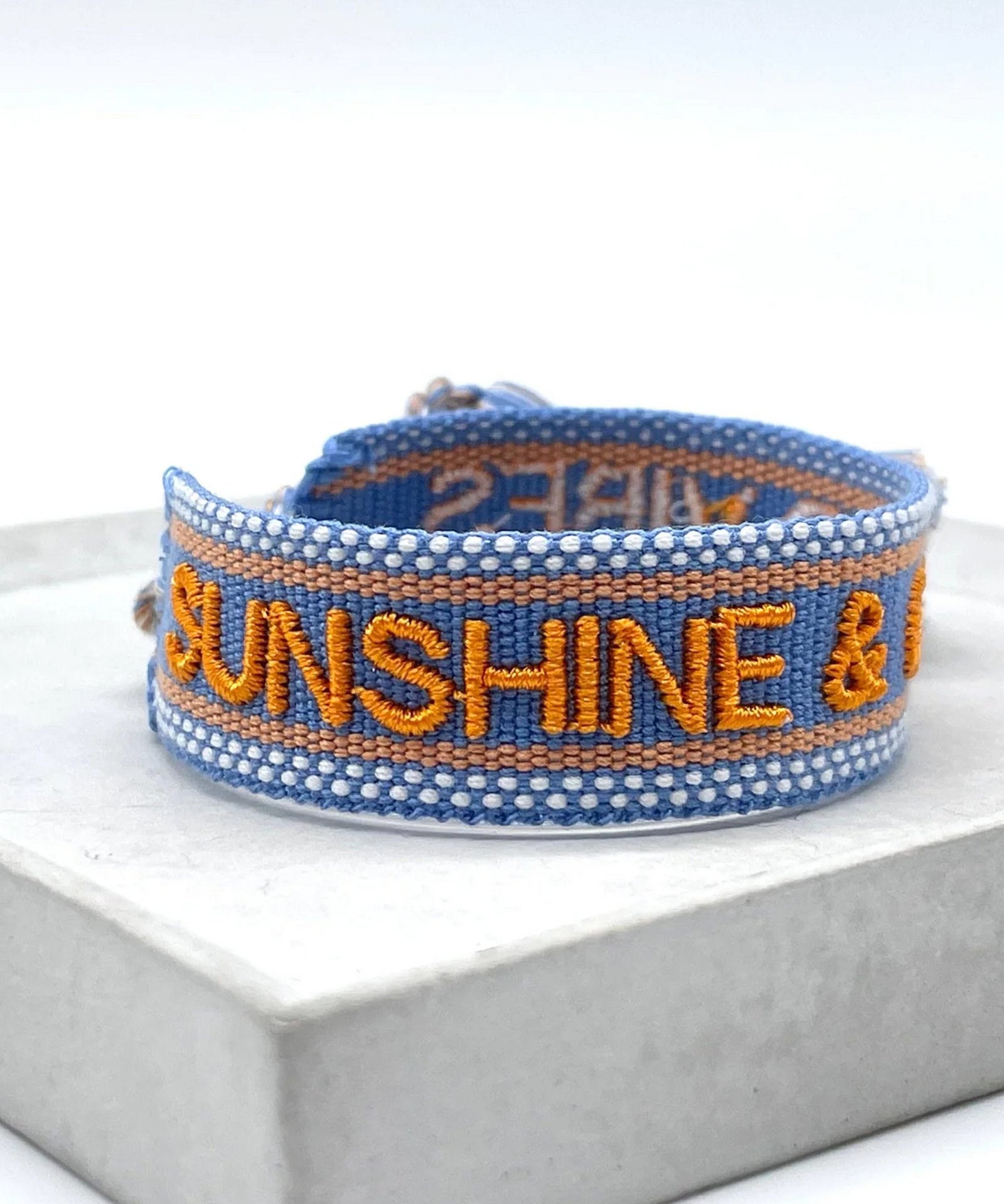 Close-up of adjustable blue and orange woven friendship bracelet featuring embroidered "SUNSHINE & GOOD VIBES" lettering and sliding knot tassel cords.