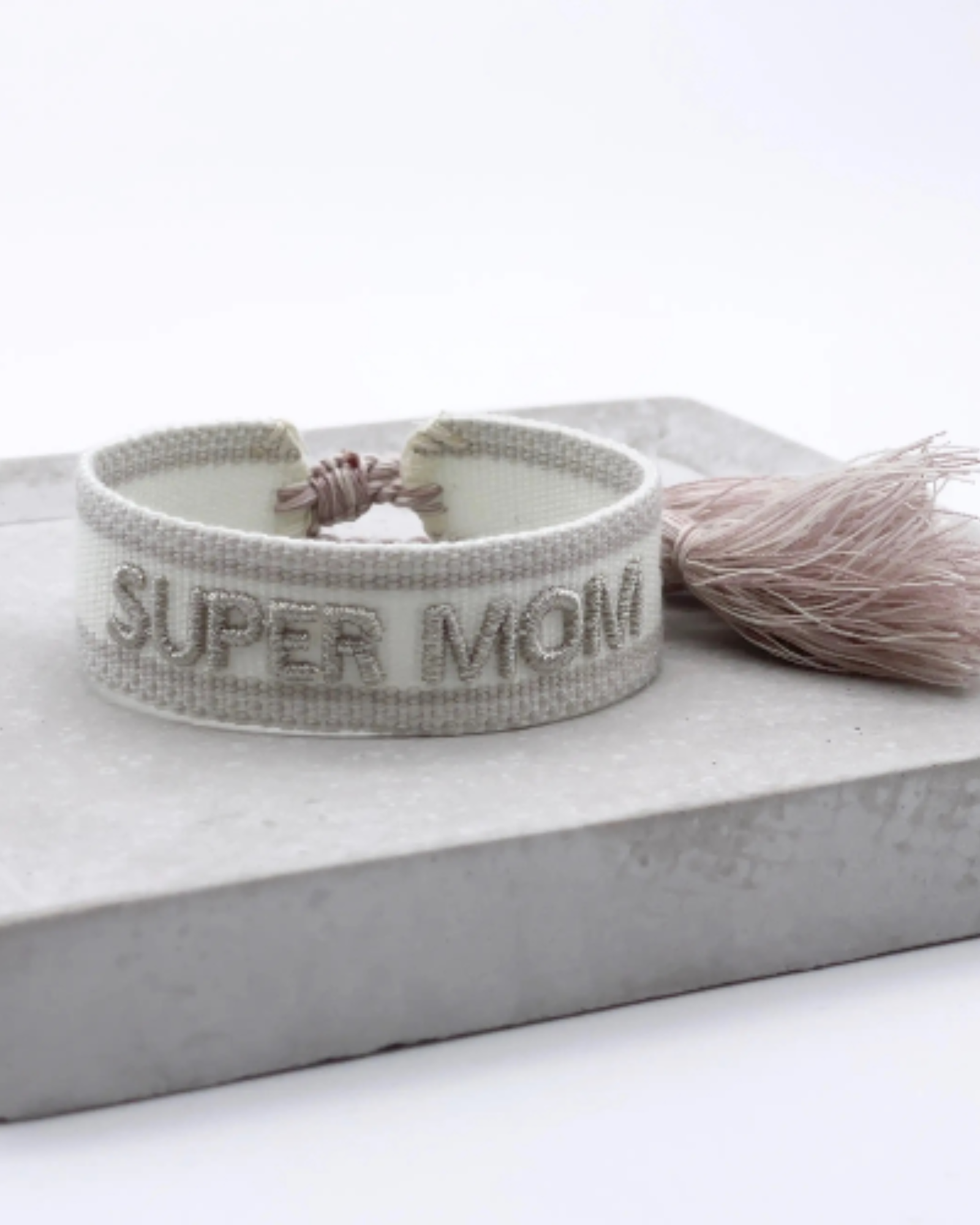 Close-up of adjustable white woven friendship bracelet with silver embroidered "SUPER MOM" lettering and sliding knot tassel cords.