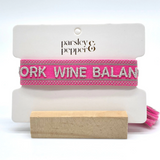 Close-up of adjustable pink woven bracelet with silver gray embroidered “WORK WINE BALANCE” lettering, matching border, and sliding knot tassel cords.