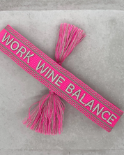 Close-up of adjustable pink woven bracelet with silver gray embroidered “WORK WINE BALANCE” lettering, matching border, and sliding knot tassel cords.