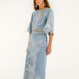 Front view of a model in a studio wearing the Farm Rio denim Richelieu wavy hem midi skirt paired with a matching embroidered top.