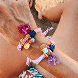 Colourful GOUPI bandana bracelet with a lilac glass crab charm on a person's wrist on summer vacations on a beach. 