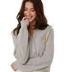 A close-up front view of a smiling woman wearing the classic grey Keila summer fine knit pullover, showcasing the partial buttoned neckline and yellow embroidered collegiate patch.
