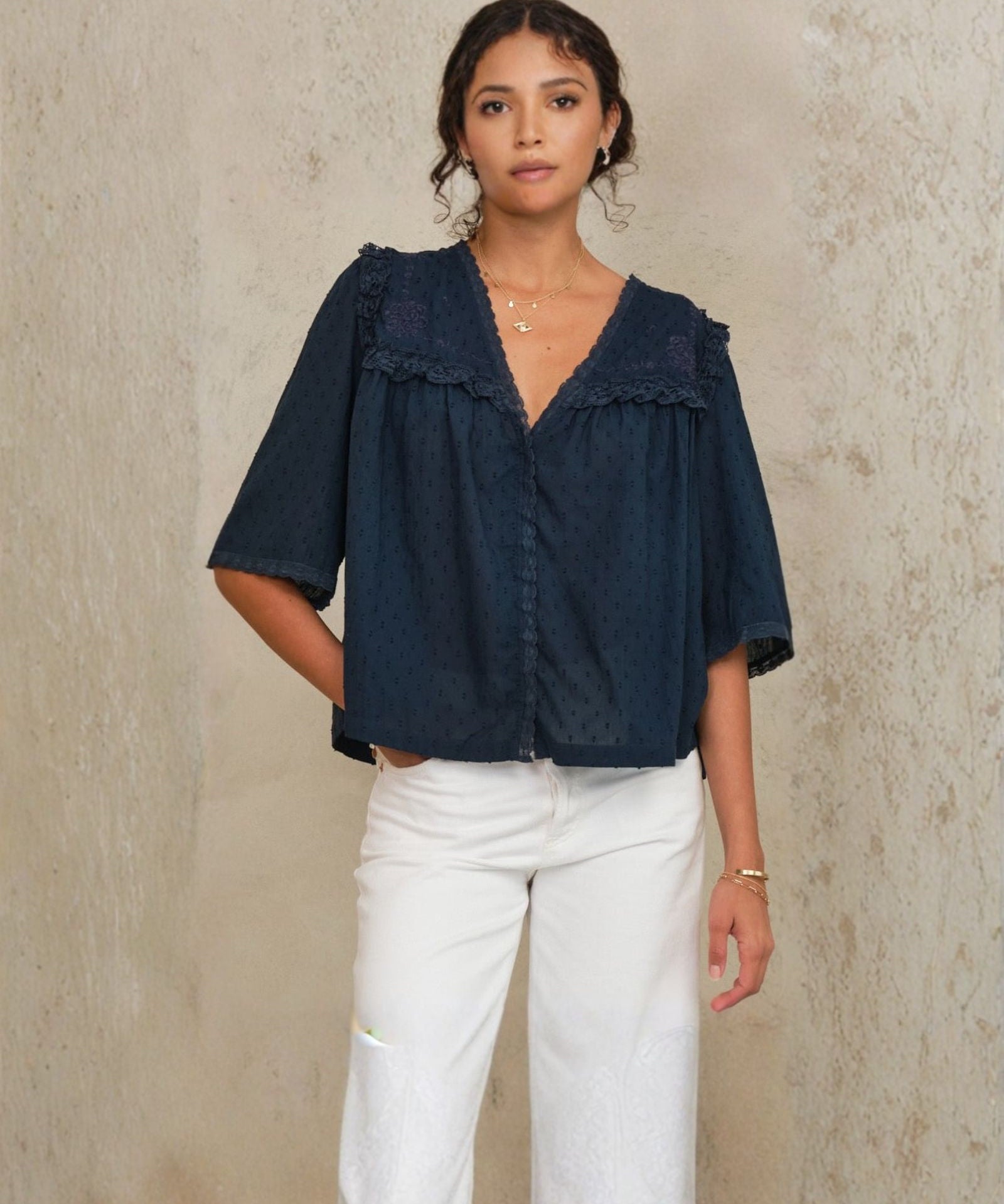 Model wearing Mabe Apparel navy embroidered cotton blouse with white trousers and jewelry.