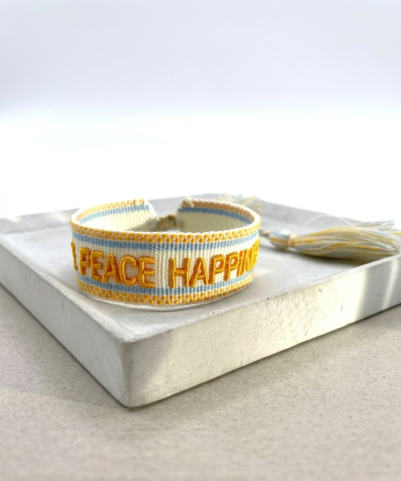 Close-up of adjustable sand-colored woven bracelet with yellow embroidered “LOVE PEACE HAPPINESS” lettering, yellow and light blue borders, and sliding knot tassel cords.