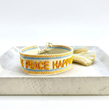 Cream woven Parsley & Pepper bracelet with mustard-yellow “LOVE PEACE HAPPINESS” embroidery, yellow and light blue borders and tasselled sliding knot.