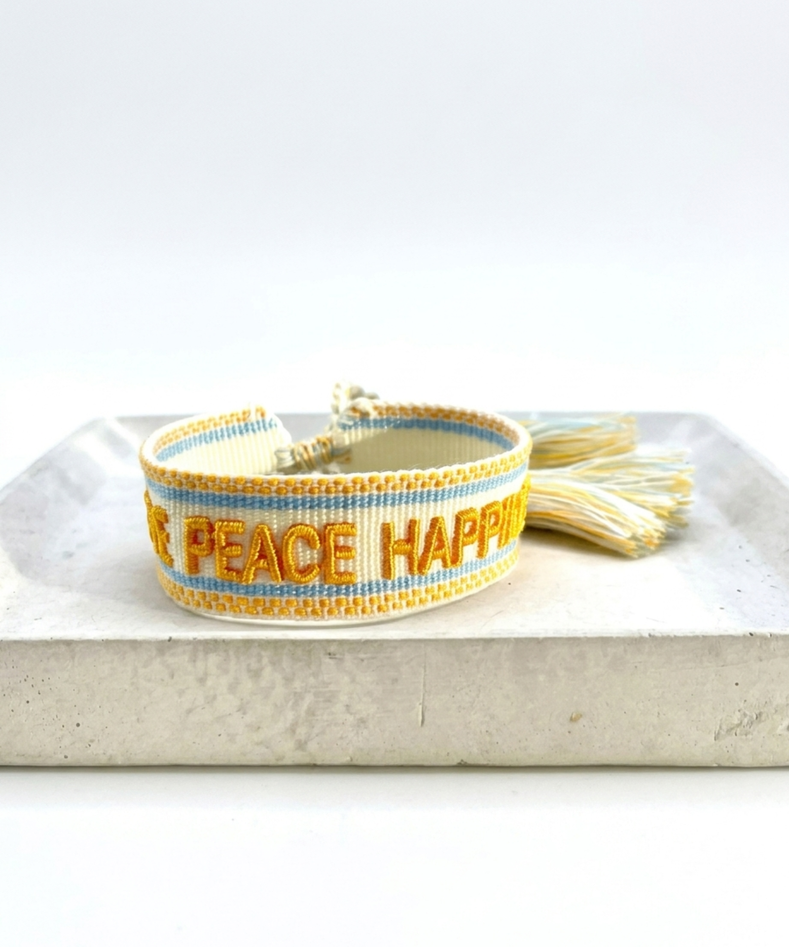 Close-up of adjustable sand-colored woven bracelet with yellow embroidered “LOVE PEACE HAPPINESS” lettering, yellow and light blue borders, and sliding knot tassel cords.