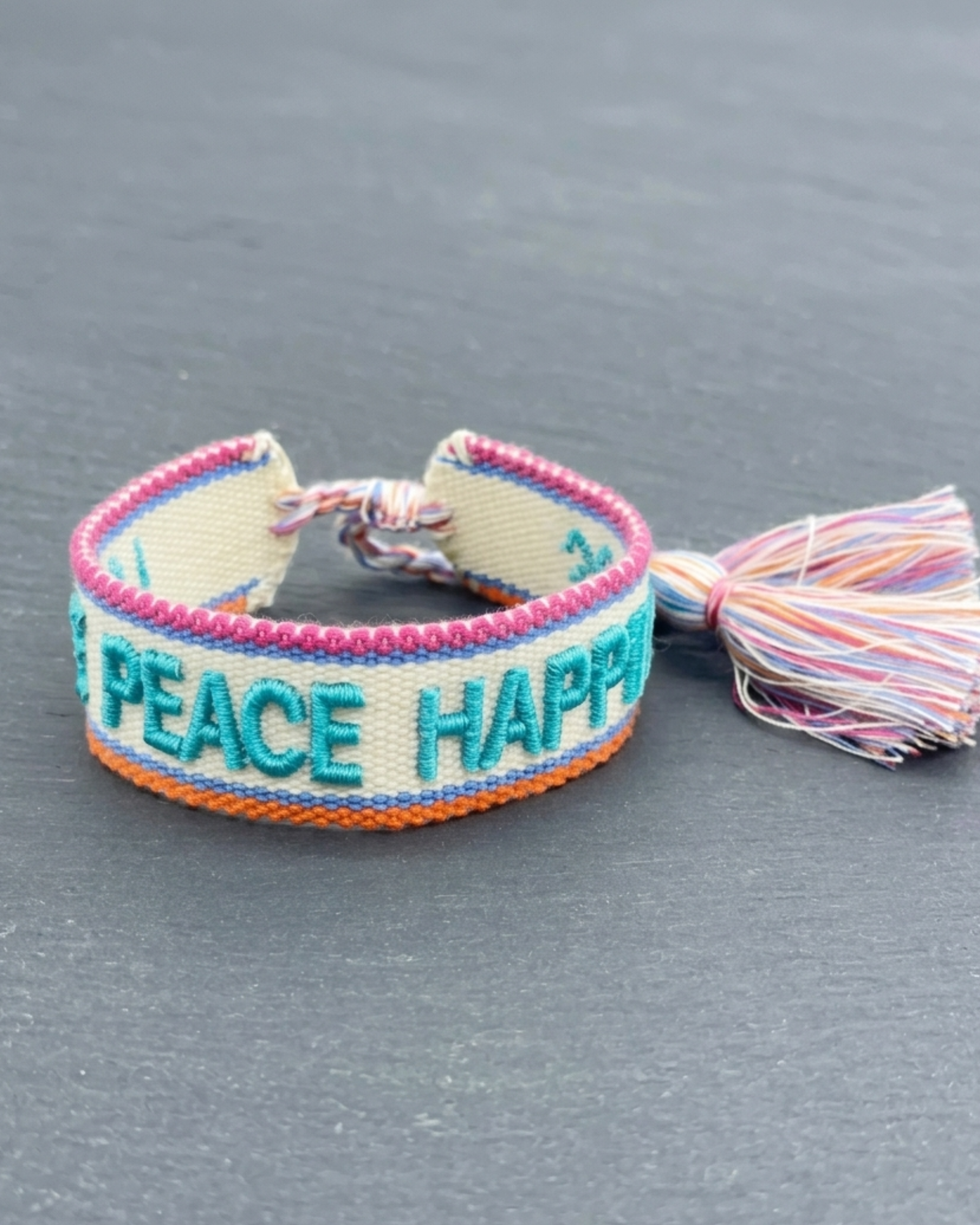 Close-up of adjustable woven turquoise friendship bracelet with embroidered “LOVE PEACE HAPPINESS” lettering and sliding knot tassel cords.