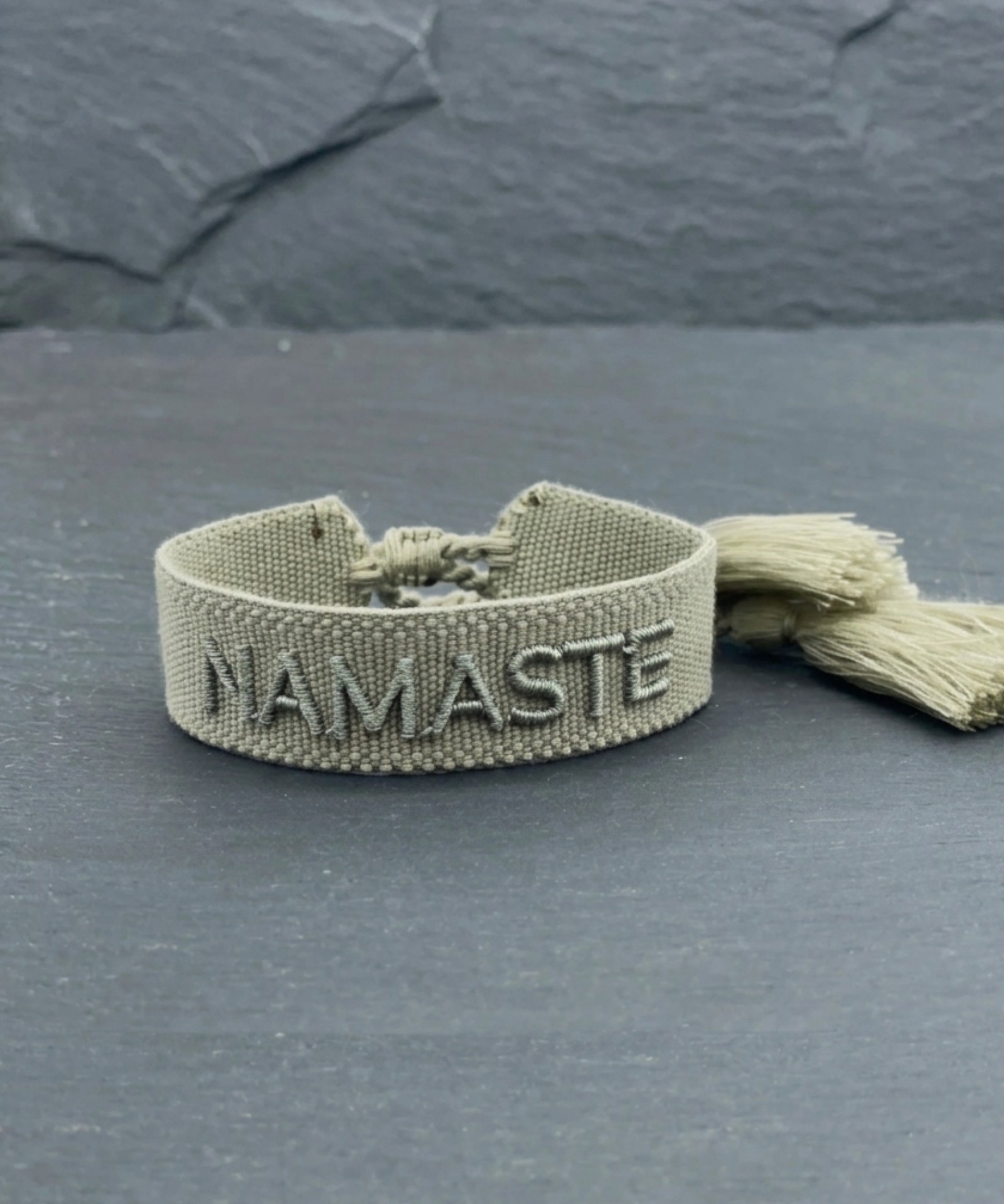 Close-up of adjustable sage eucalyptus woven bracelet with tone-on-tone embroidered “NAMASTE” lettering and sliding knot tassel cords.
