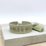 Close-up of adjustable platinum sage woven bracelet with tone-on-tone embroidered “SMILE & SHINE” lettering and sliding knot tassel cords.