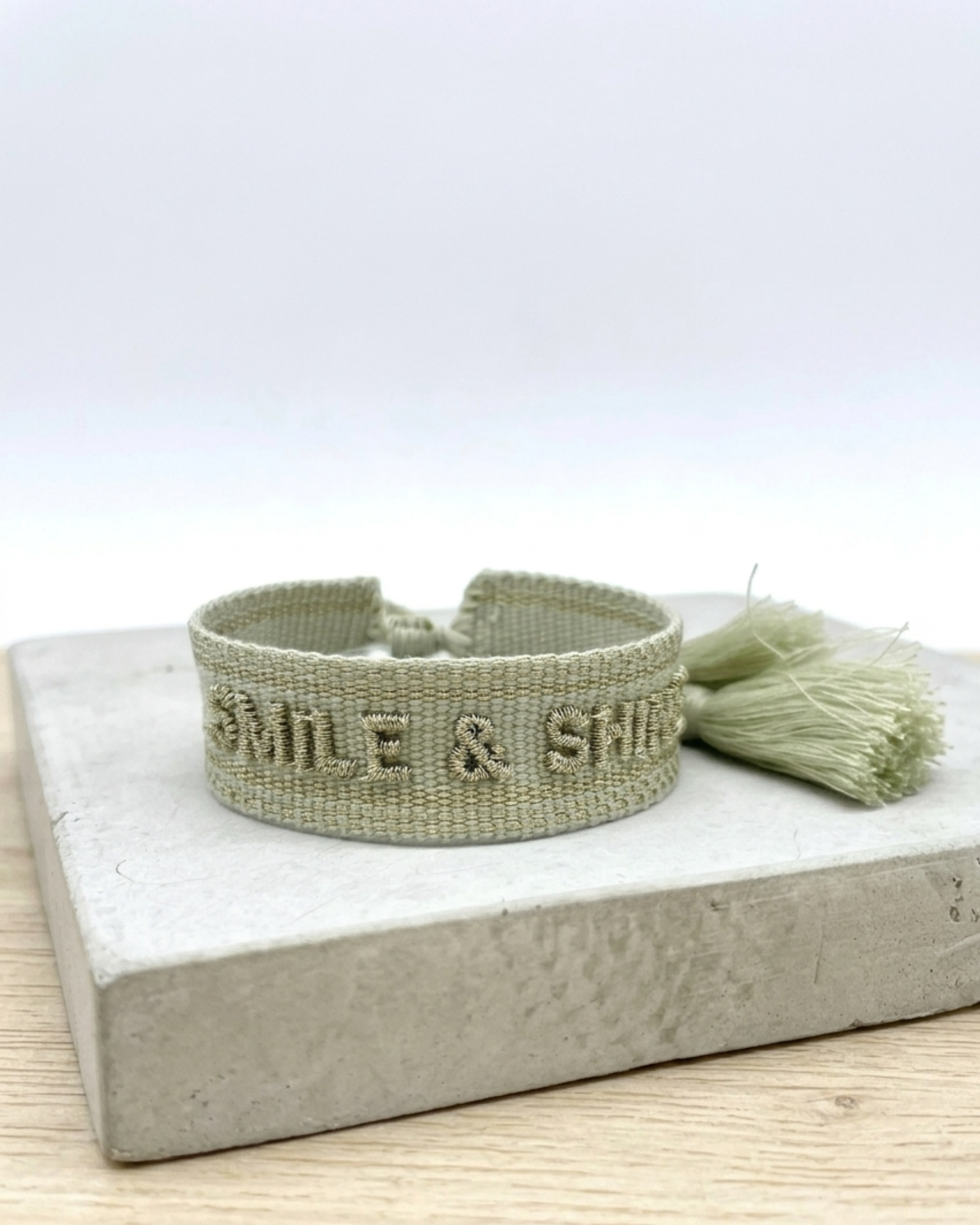 Close-up of adjustable platinum sage woven bracelet with tone-on-tone embroidered “SMILE & SHINE” lettering and sliding knot tassel cords.