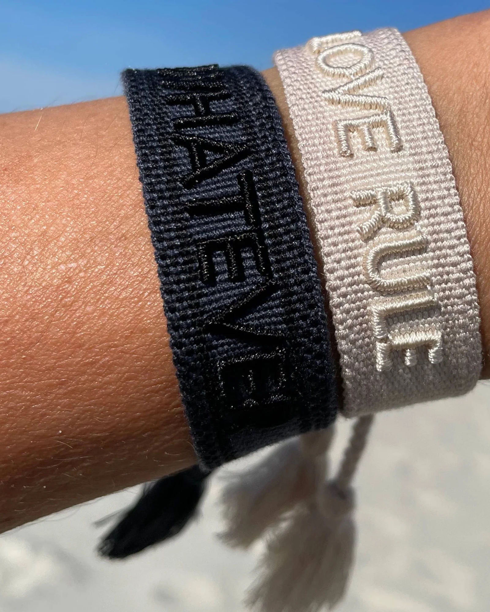 Close-up of adjustable dark grey woven bracelet with black embroidered “WHATEVER” lettering, black border, and sliding knot tassel cords.