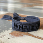 Close-up of adjustable dark grey woven bracelet with black embroidered “WHATEVER” lettering, black border, and sliding knot tassel cords.