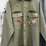 Front studio view of the khaki military shirt jacket hanging, showcasing the two chest pockets, full button closure, and extensive collection of pins and colorful custom patches.