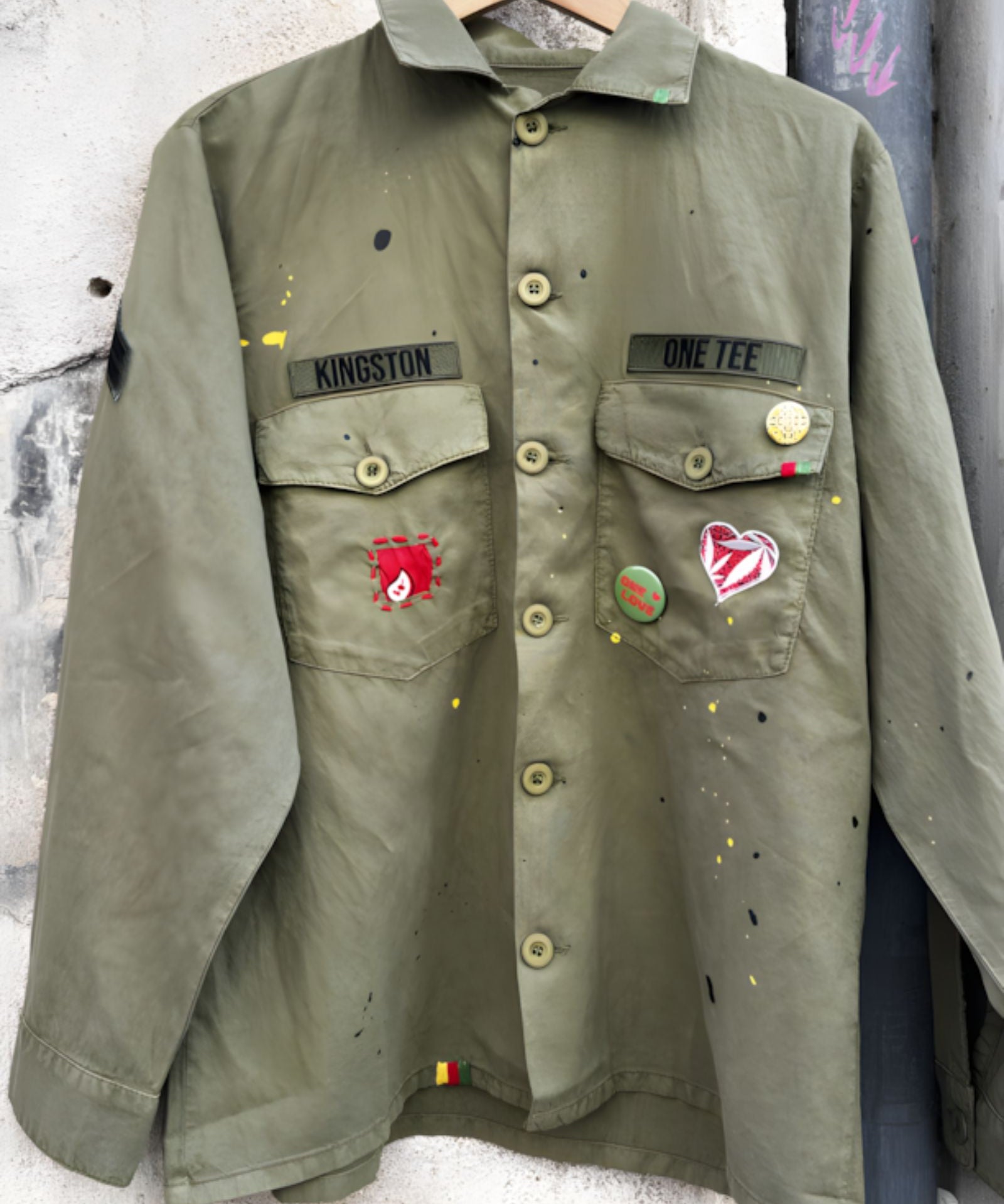 Front studio view of the khaki military shirt jacket hanging, showcasing the two chest pockets, full button closure, and extensive collection of pins and colorful custom patches.
