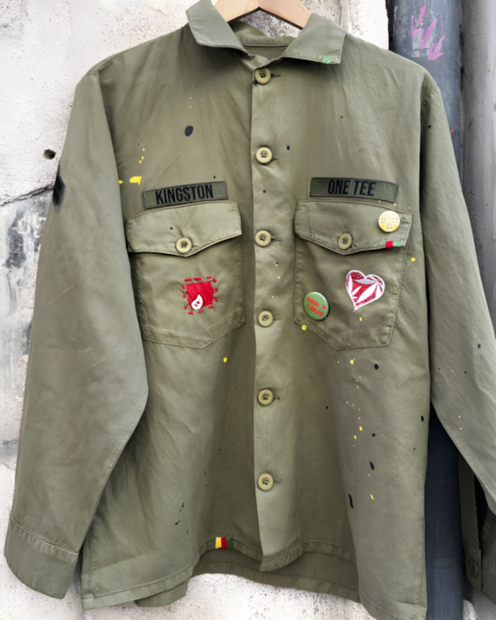 Front studio view of the khaki military shirt jacket hanging, showcasing the two chest pockets, full button closure, and extensive collection of pins and colorful custom patches.