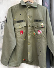 Front studio view of the khaki military shirt jacket hanging, showcasing the two chest pockets, full button closure, and extensive collection of pins and colorful custom patches.