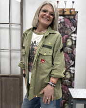A smiling woman wearing the khaki green Rita military patch shirt jacket over a graphic t-shirt, displaying its straight fit and unique front patch details.