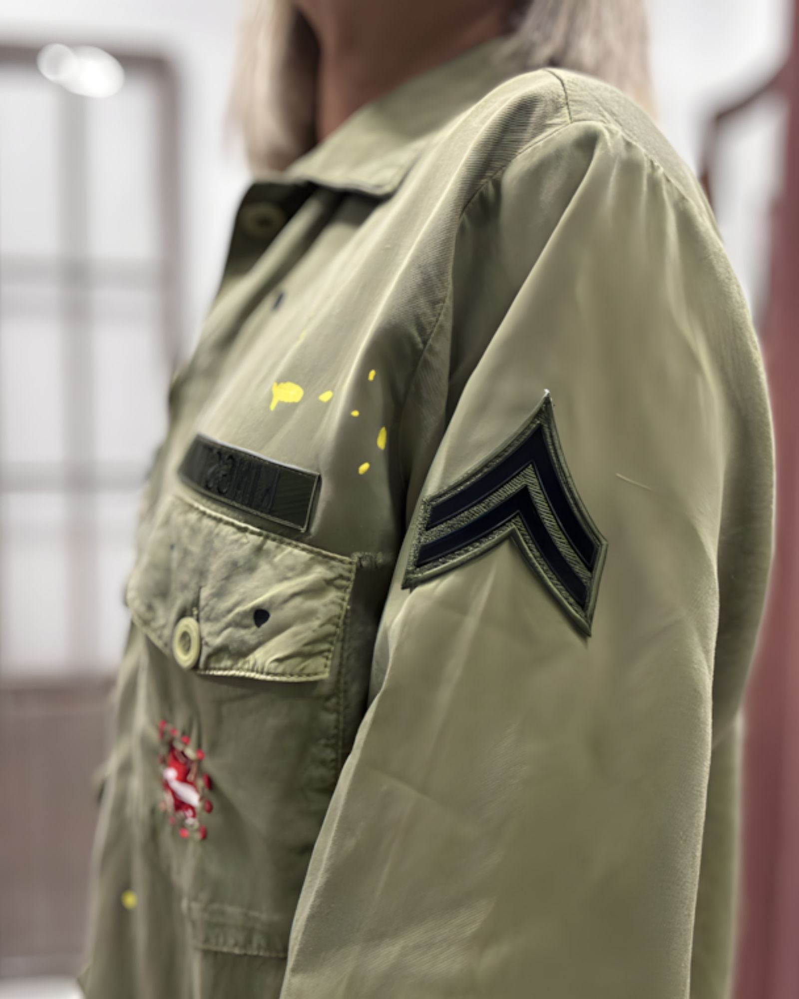 Close-up side view of the Rita military jacket, highlighting the classic khaki fabric texture and the black chevron patch embroidered on the sleeve.
