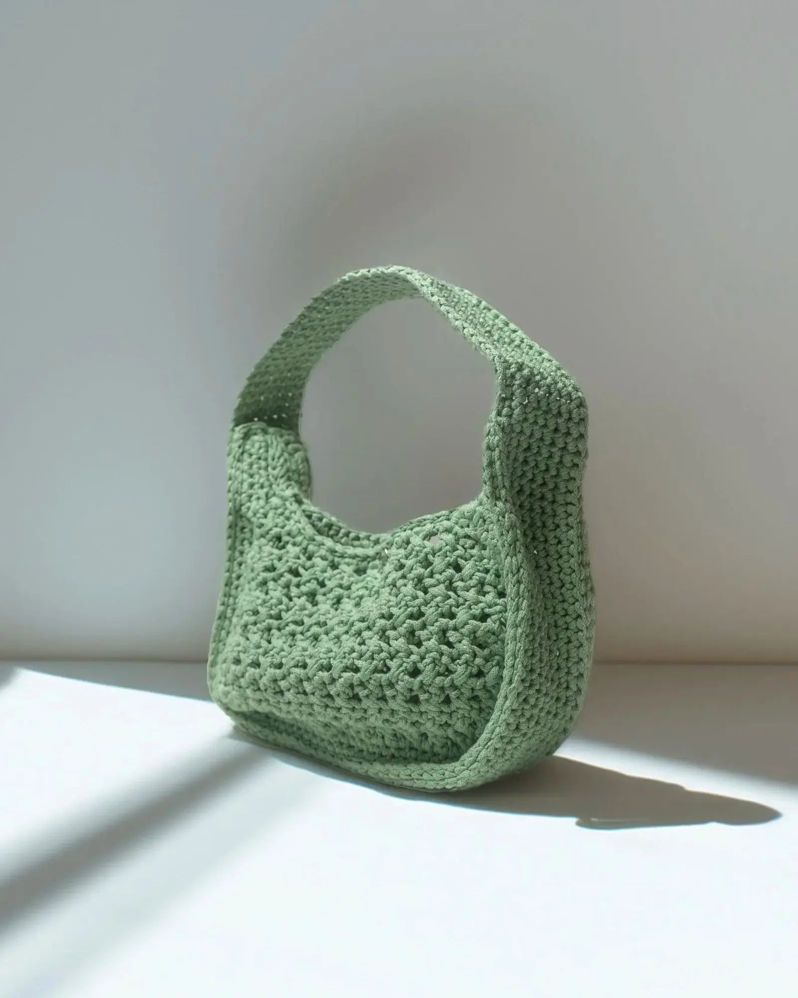 Riviera Lili handcrafted crochet bag in sage green standing in natural light with shadow — Atelier Bagatelle Switzerland