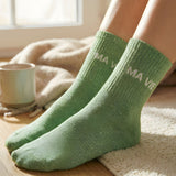 Close-up shot of the model's feet on a wooden floor, highlighting the soft texture and the white jacquard MA VIE inscription on the green glitter socks.