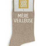 Elegant beige glitter message socks with a white jacquard MÈRE VEILLEUSE inscription at the ankle, presented in their original Woom paper packaging.