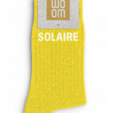 Vibrant yellow glitter message socks with a white jacquard SOLAIRE inscription at the ankle, presented in their original Woom paper packaging.